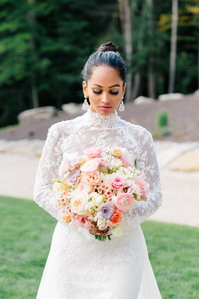 Newport Vineyards Wedding | Caprio Captured Photography Light and Airy Wedding Photography | A bride in a lacy white gown holds a colorful bouquet of pink and peach flowers. She stands outdoors on grass, with a serene, thoughtful expression.