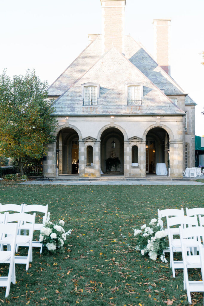 Glen Manor House Wedding Cost | Caprio Captured Photography Light and Airy Wedding Photography | Elegant stone building with arched doorways and a slate roof, set in a grassy yard lined with white chairs and floral arrangements, suggesting a wedding.
