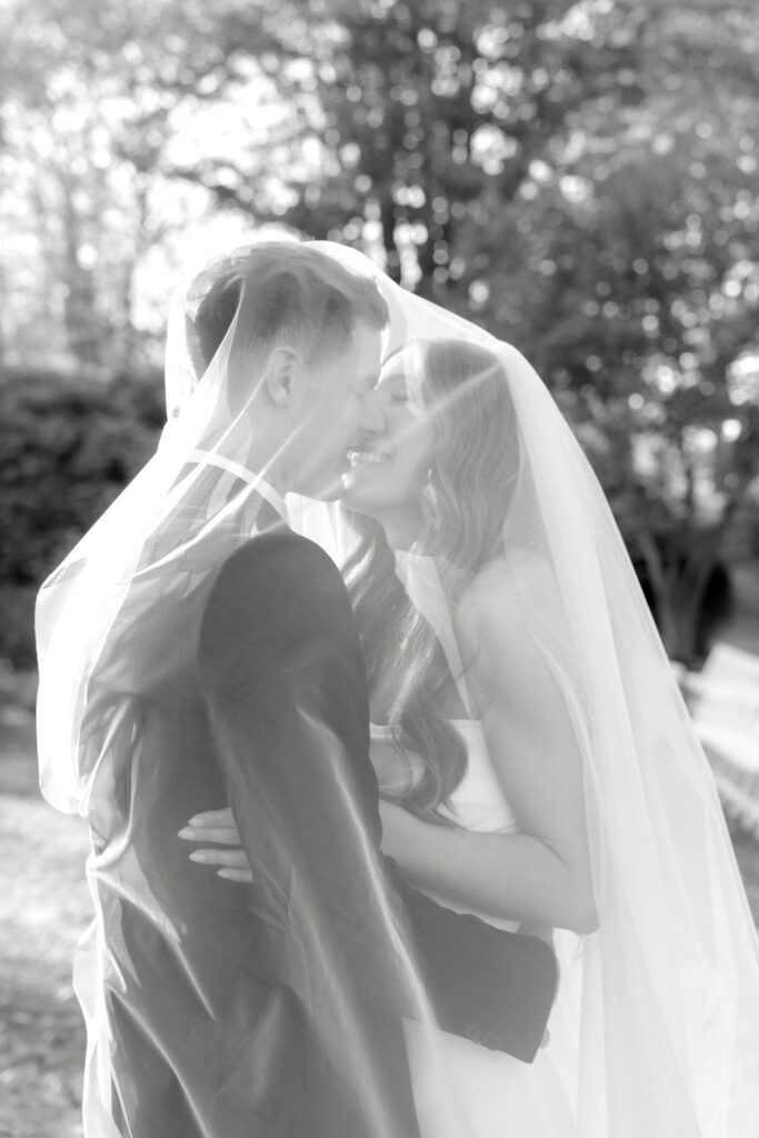 Glen Manor House Wedding Cost | Caprio Captured Photography Light and Airy Wedding Photography | A couple embraces under sheer veils, sharing a joyful moment in a sunlit outdoor setting. The black-and-white photo exudes romance and happiness.
