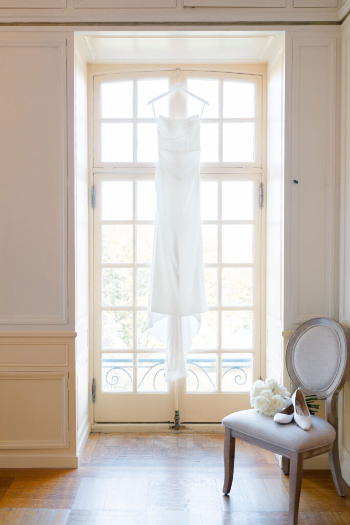 Glen Manor House Wedding Cost | Caprio Captured Photography Light and Airy Wedding Photography | Elegant wedding dress hangs in bright window. Nearby, a chair holds white heels and flowers, conveying a serene, anticipatory atmosphere.