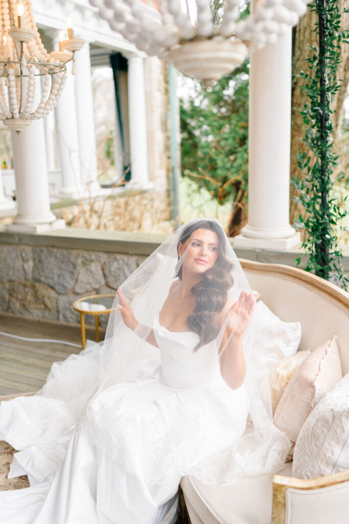 Newport RI Mansion Wedding | Caprio Captured Photography Light and Airy Wedding Photography | Bride in white dress and veil sits on a cream sofa under chandeliers on a veranda with stone columns, surrounded by greenery and elegant decor.