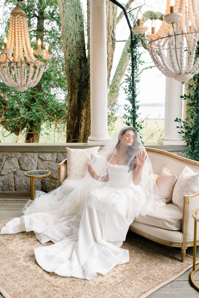 Newport Mansions Weddings Cost | Caprio Captured Photography Light and Airy Wedding Photography | Bride in elegant white gown and veil sits on a vintage sofa under chandeliers, surrounded by greenery. She appears serene and contemplative.