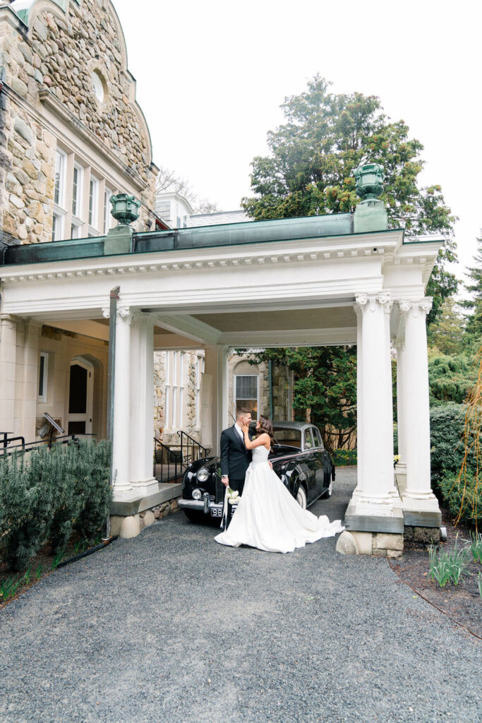 Newport RI Mansion Wedding | Caprio Captured Photography Light and Airy Wedding Photography | Bride and groom embrace under a grand stone archway beside a classic black car. The setting is elegant, with lush greenery and an ornate building façade.