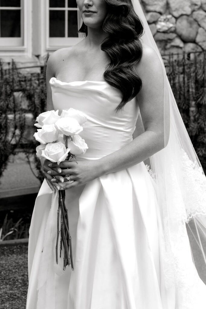 Shining Tides Weddings | Caprio Captured Photography Light and Airy Wedding Photography | Bride in a strapless gown holding white roses. Her long, wavy hair and veil accentuate her elegant look. A stone wall and window form the backdrop.