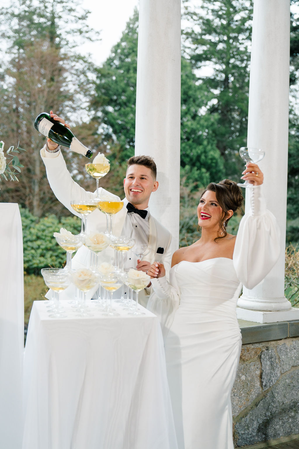 Newport RI Mansion Wedding | Caprio Captured Photography Light and Airy Wedding Photography | A couple in elegant wedding attire is joyfully pouring champagne into a tiered tower of glasses. The setting is outdoor with lush greenery.