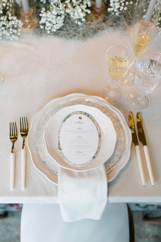 Belle Mer Wedding Price | Caprio Captured Photography Light and Airy Wedding Photography | Elegant table setting with white and gold cutlery flanking decorative plates. A menu sits atop, with glasses of champagne nearby, exuding sophistication.