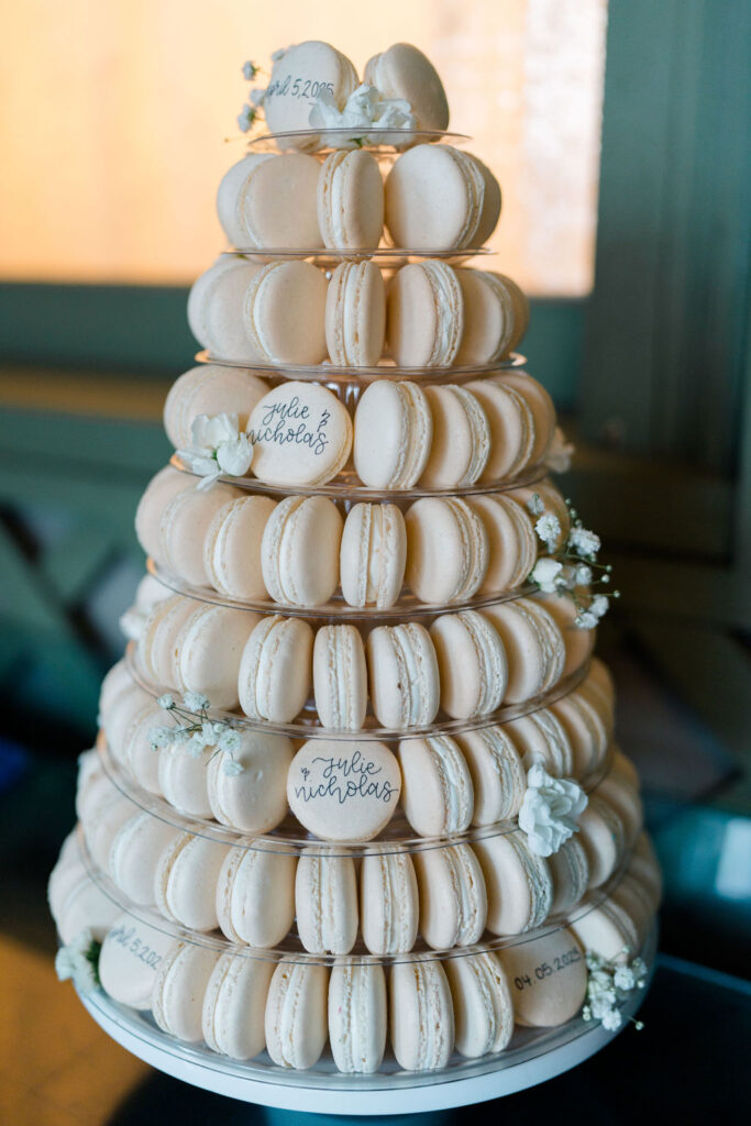 Shining Tides Weddings | Caprio Captured Photography Light and Airy Wedding Photography | A pyramid of ivory macarons on a tiered stand adorned with white flowers. Some macarons display handwritten names and dates, evoking a wedding theme.