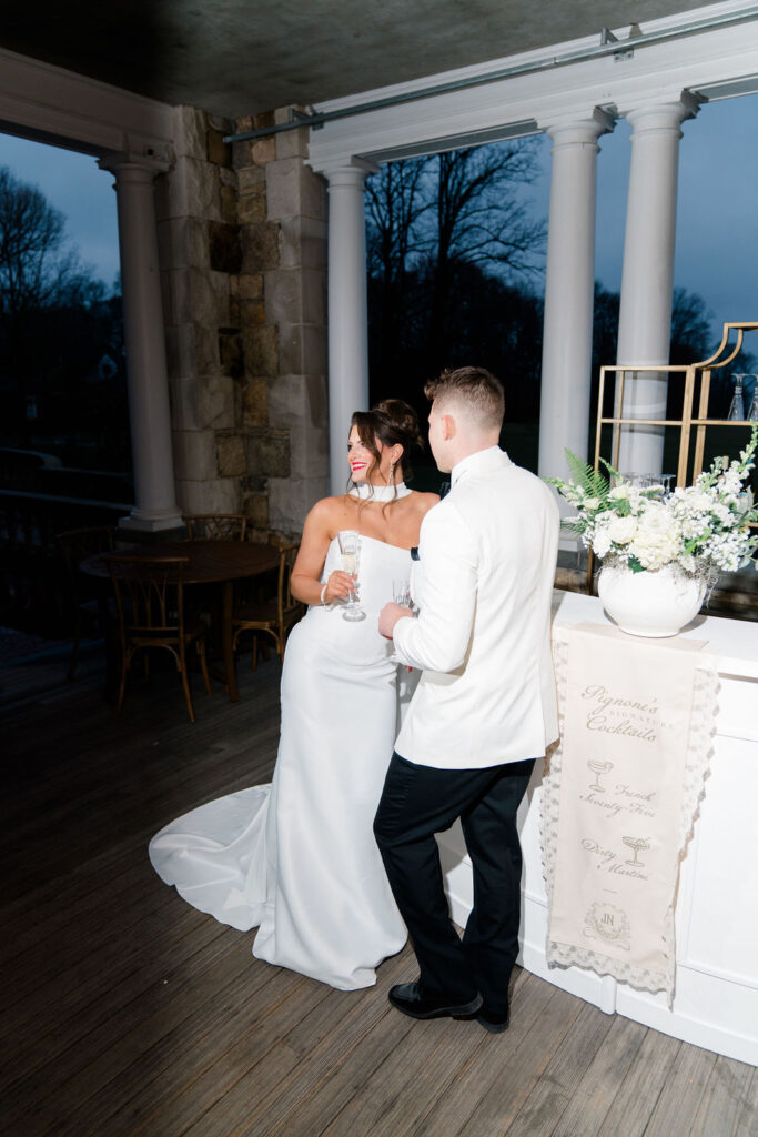Newport RI Mansion Wedding | Caprio Captured Photography Light and Airy Wedding Photography | A bride in a white dress and groom in a white jacket stand on a terrace, holding champagne glasses. They smile at each other with elegant decor around.
