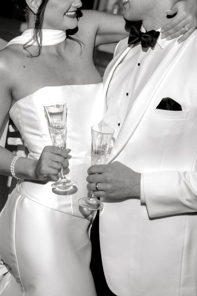 Newport RI Mansion Wedding | Caprio Captured Photography Light and Airy Wedding Photography | A couple in formal attire, holding champagne flutes, smiling and embracing. The black and white image conveys elegance and celebration.