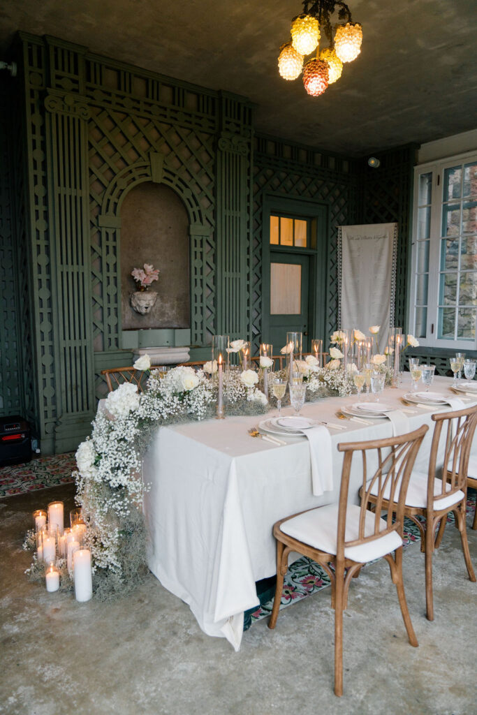 Newport RI Mansion Wedding | Caprio Captured Photography Light and Airy Wedding Photography | Elegant dining setup with a white tablecloth, candles, and floral centerpiece on a table set for six. Ornate green wall and hanging lights add charm.