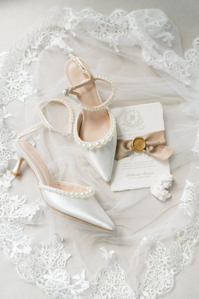 Shining Tides Weddings | Caprio Captured Photography Light and Airy Wedding Photography | Elegant bridal theme with white satin pearl-embellished heels on lace fabric, alongside an ornate invitation, gold seal, and taupe ribbon.