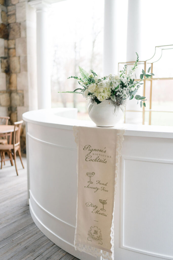Shining Tides Weddings | Caprio Captured Photography Light and Airy Wedding Photography | A white curved bar with a flower arrangement in a vase on top. A banner hangs in front, listing drinks "French 75" and "Dirty Martini" in elegant script.