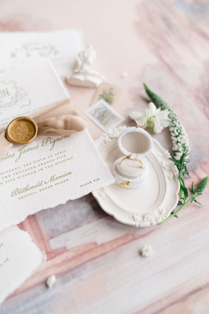 Newport RI Mansion Wedding | Caprio Captured Photography Light and Airy Wedding Photography | Elegant wedding invitation set on a soft, pastel surface. Includes a gold wax seal, vintage ring in ornate box, delicate flowers, and formal script.
