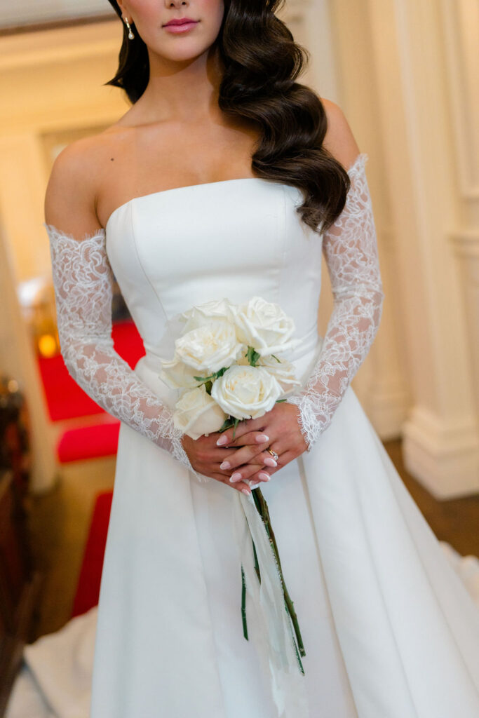 Newport RI Mansion Wedding | Caprio Captured Photography Light and Airy Wedding Photography | Bride in a white gown with lace sleeves holds a bouquet of white roses. Her hair is styled in elegant waves. The background is softly lit with a red carpet.