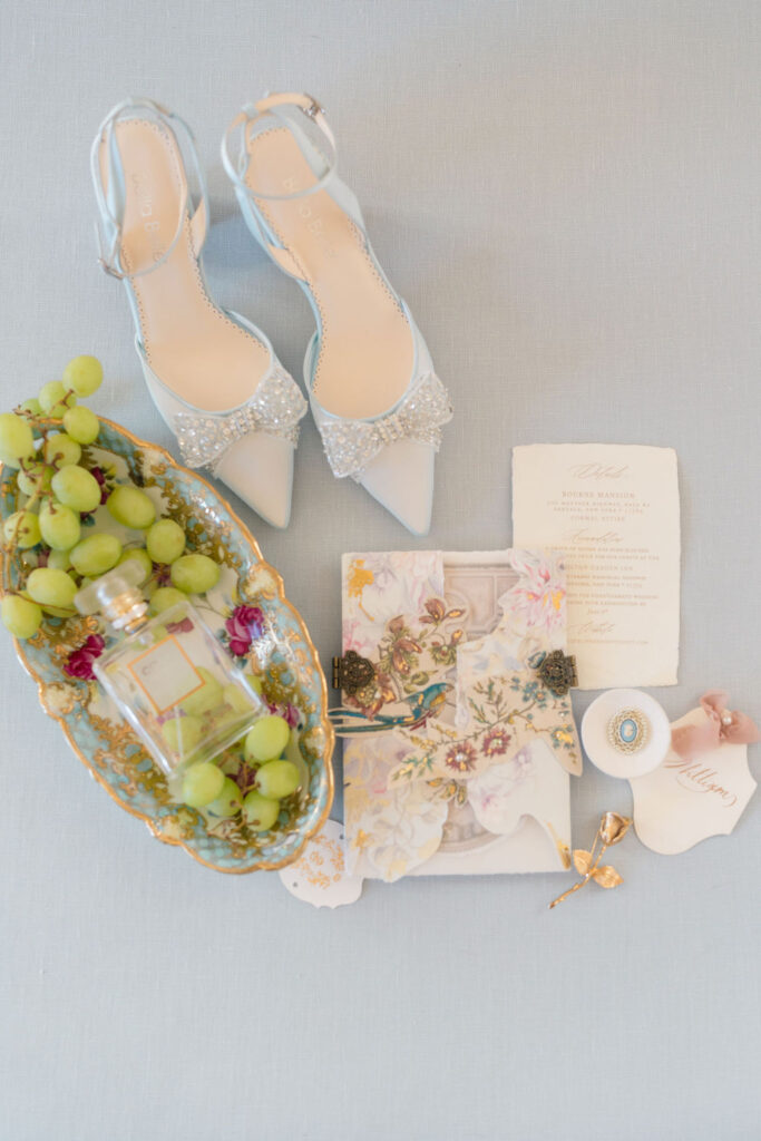 Belle Mer Wedding Price | Caprio Captured Photography Light and Airy Wedding Photography | Elegant flat lay featuring bejeweled heels, a vintage dish with green grapes and perfume, intricate invitations, and ornate earrings on a soft blue background.