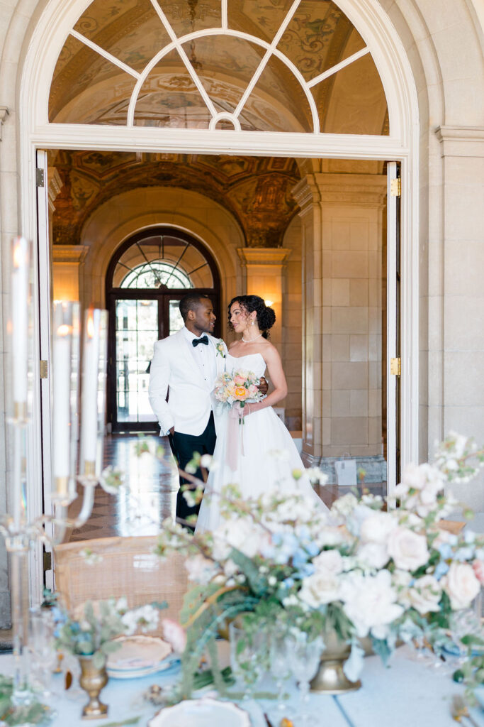 Aldrich Mansion Wedding Photos | Caprio Captured Photography Light and Airy Wedding Photography | Bride and groom in elegant attire stand in ornate doorway, smiling at each other. A decorated table with flowers and candles is in the foreground.