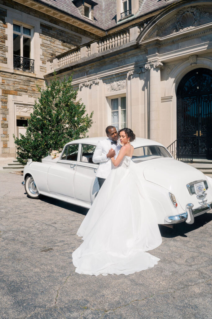 Newport Wedding Photography | Caprio Captured Photography Light and Airy Wedding Photography | Bride and groom in elegant wedding attire stand lovingly beside a vintage white car. The backdrop is a grand stone mansion under a clear sky.