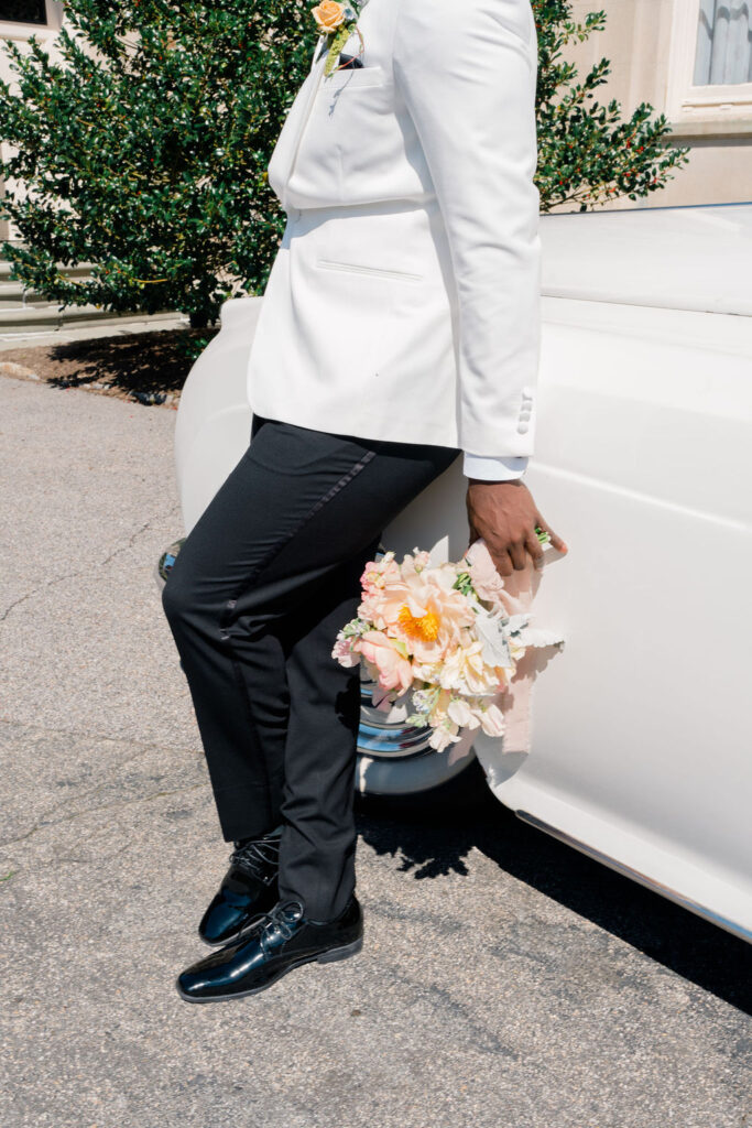 Breakers Mansion Newport Rhode Island | Caprio Captured Photography Light and Airy Wedding Photography | Man in a white suit jacket and black pants leans on a vintage white car, holding a bouquet of pastel flowers. The mood is elegant and serene.