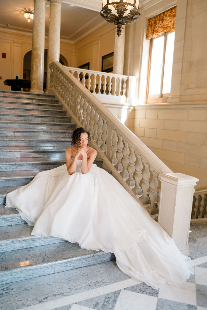 Newport Wedding Photography | Caprio Captured Photography Light and Airy Wedding Photography | Bride in an elegant white gown sits on a grand marble staircase with ornate railings, hands gently clasped; soft lighting adds a serene ambiance.