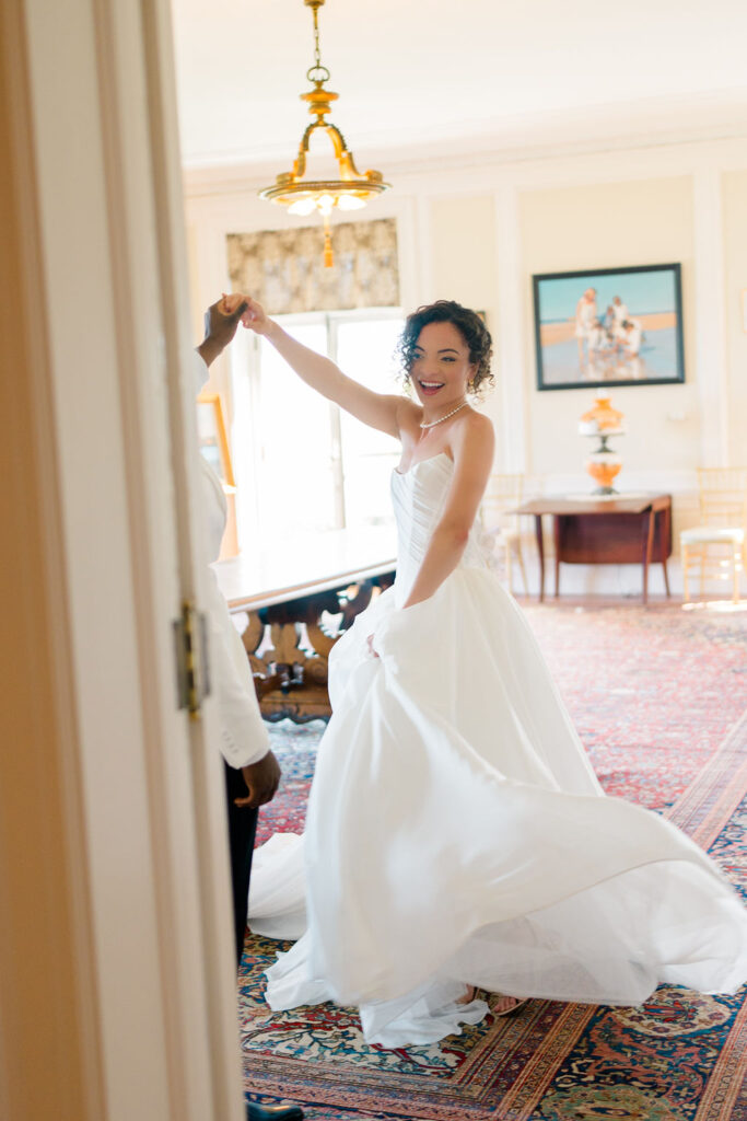 Newport Mansions Weddings Cost | Caprio Captured Photography Light and Airy Wedding Photography | Bride in a white gown twirling joyfully with a partner in a bright, elegant room adorned with vintage decor and warm lighting.