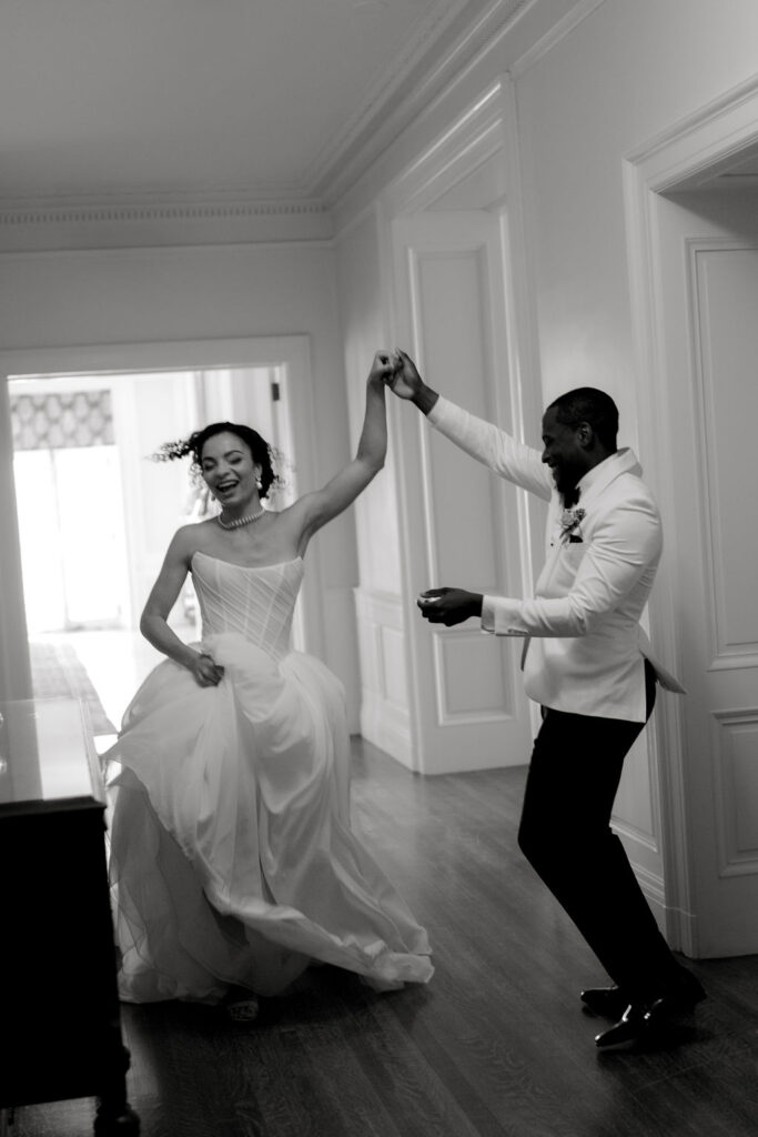 Newport Wedding Photography | Caprio Captured Photography Light and Airy Wedding Photography | A joyful bride and groom dance inside a hallway. The bride holds her dress, smiling brightly, while the groom twirls her with a beaming smile.