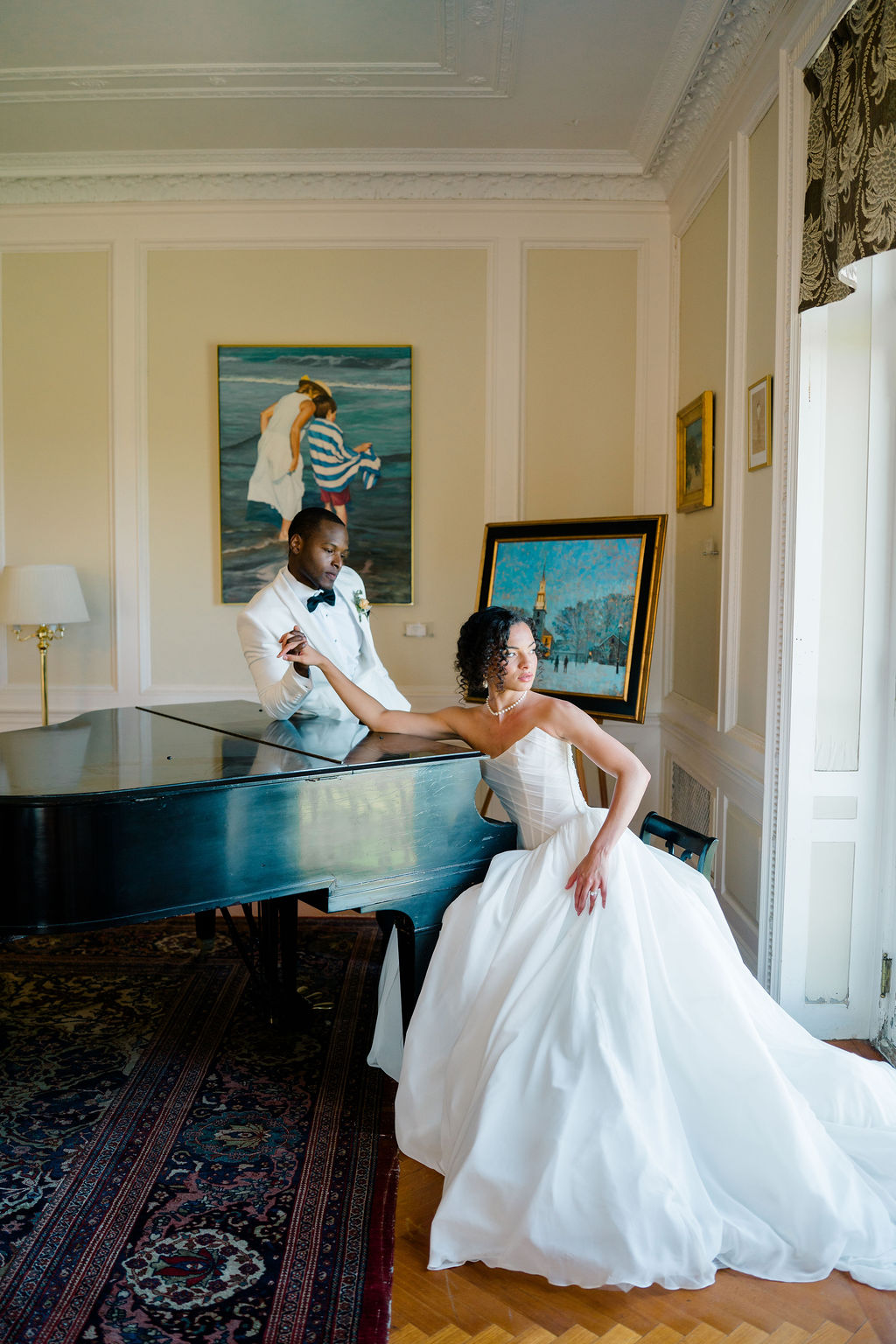 Newport Wedding Photography | Caprio Captured Photography Light and Airy Wedding Photography | A couple in wedding attire poses near a grand piano in an elegant room with paintings and soft lighting. The scene conveys romance and sophistication.