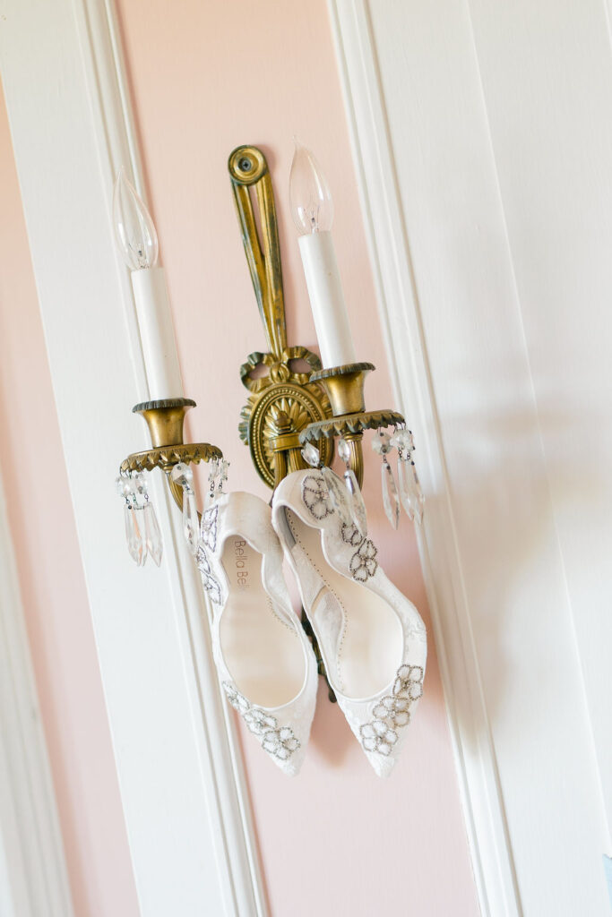 Newport Wedding Photography | Caprio Captured Photography Light and Airy Wedding Photography | Elegant white heels with floral lace rest on a vintage brass wall sconce. The background features soft pink and white paneling, evoking a romantic tone.