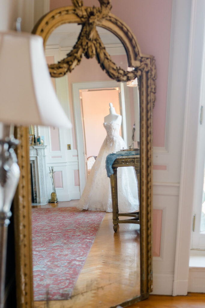 Newport Mansions Weddings Cost | Caprio Captured Photography Light and Airy Wedding Photography | Reflection of an elegant white bridal gown on a mannequin in an ornate mirror against a pink wall, creating a romantic and vintage ambiance.