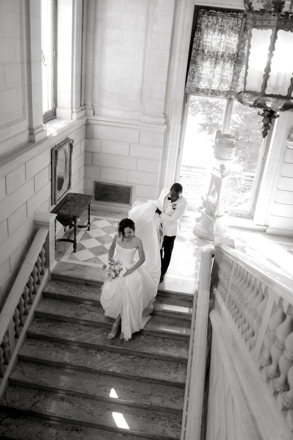 Newport Mansions Weddings Cost | Caprio Captured Photography Light and Airy Wedding Photography | Bride and groom in formal attire ascend elegant marble staircase. Bride carries bouquet and groom holds her train, with soft light filtering through large windows.