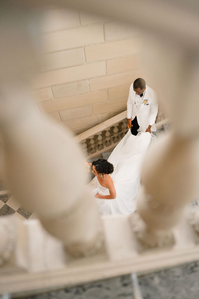 Newport Wedding Photography | Caprio Captured Photography Light and Airy Wedding Photography | Bride and groom descending a marble staircase, framed by balusters. She's in a flowing white gown, and he's in a white suit, exuding elegance.