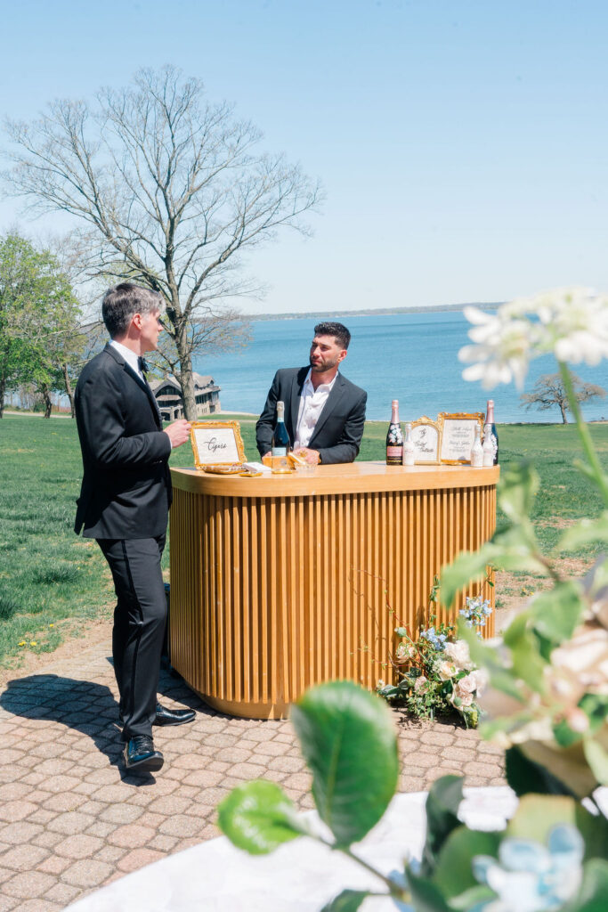 Waterfront Wedding Venues Rhode Island | Caprio Captured Photography Light and Airy Wedding Photography | Two men in suits stand at an outdoor bar adorned with drinks and signs, set against a scenic lake view and trees, evoking a relaxed, elegant vibe.