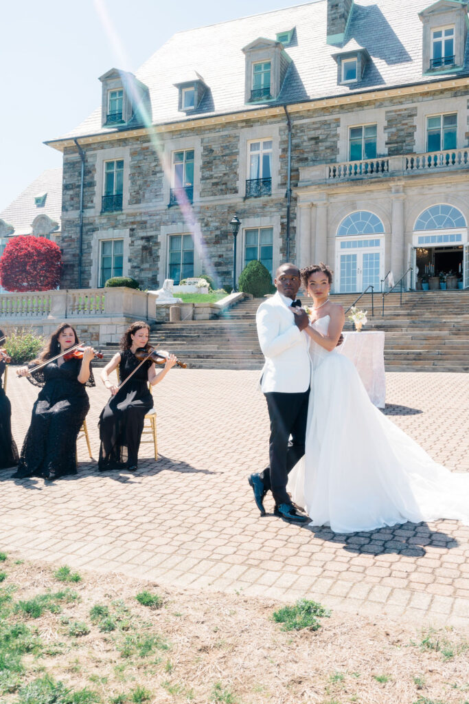 Newport Mansions Weddings Cost | Caprio Captured Photography Light and Airy Wedding Photography | A bride and groom dance outdoors before a grand stone mansion. A string quartet in black dresses plays nearby. The scene is elegant and joyful.