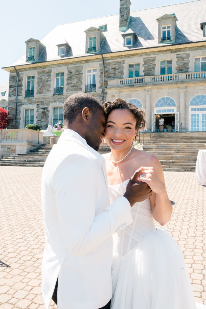 Newport Wedding Photography | Caprio Captured Photography Light and Airy Wedding Photography | A joyful couple in wedding attire, dancing and smiling outside a grand stone mansion with stairs and large windows, under a clear blue sky.