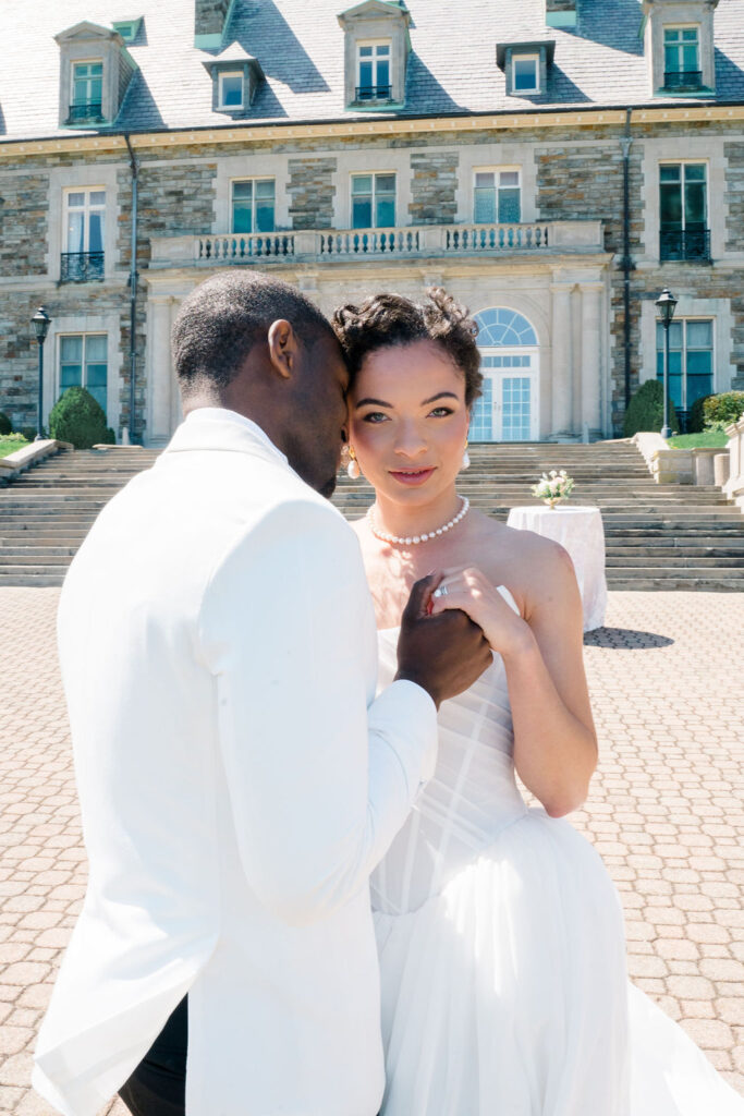Newport Mansions Weddings Cost | Caprio Captured Photography Light and Airy Wedding Photography | A couple embraces in elegant attire in front of a grand mansion. The man whispers to the woman, who smiles gently, conveying warmth and love.