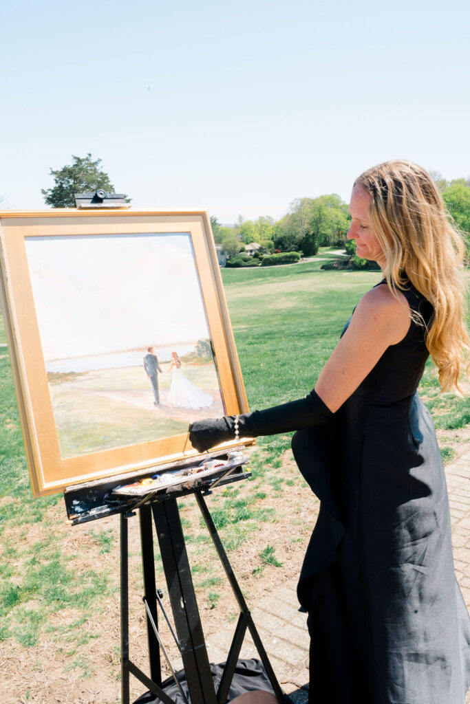 Newport Wedding Photography | Caprio Captured Photography Light and Airy Wedding Photography | A woman in a black dress paints a wedding scene outdoors on a sunny day. The painting shows a couple holding hands on a grassy field.
