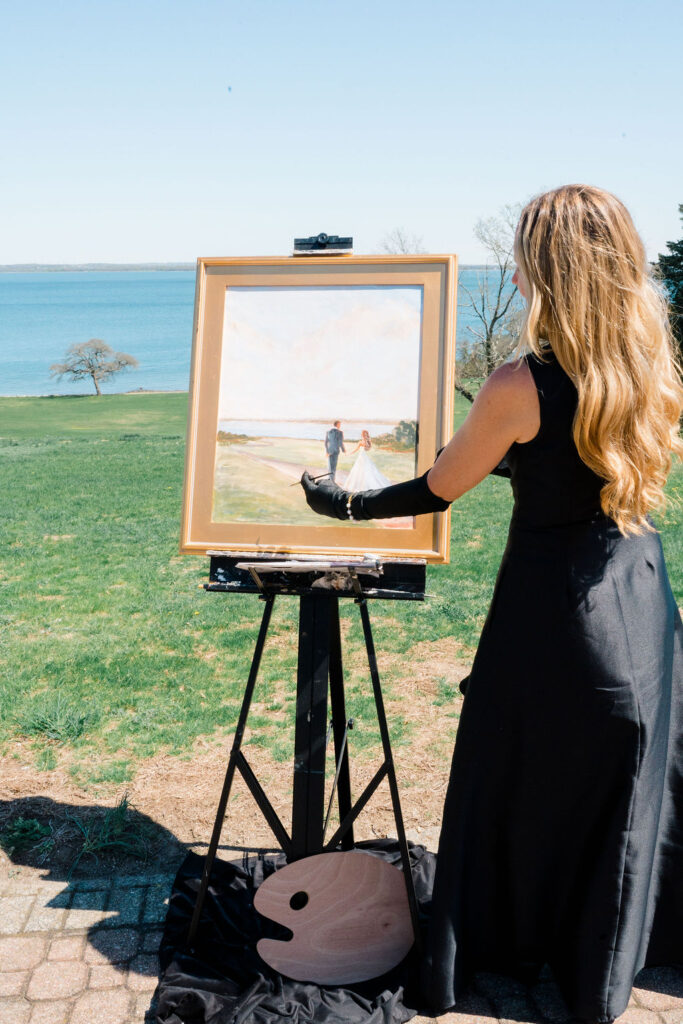 Shining Tides Weddings | Caprio Captured Photography Light and Airy Wedding Photography | A woman in a black dress paints a wedding scene on an easel outdoors, overlooking a grassy area and a serene blue lake.