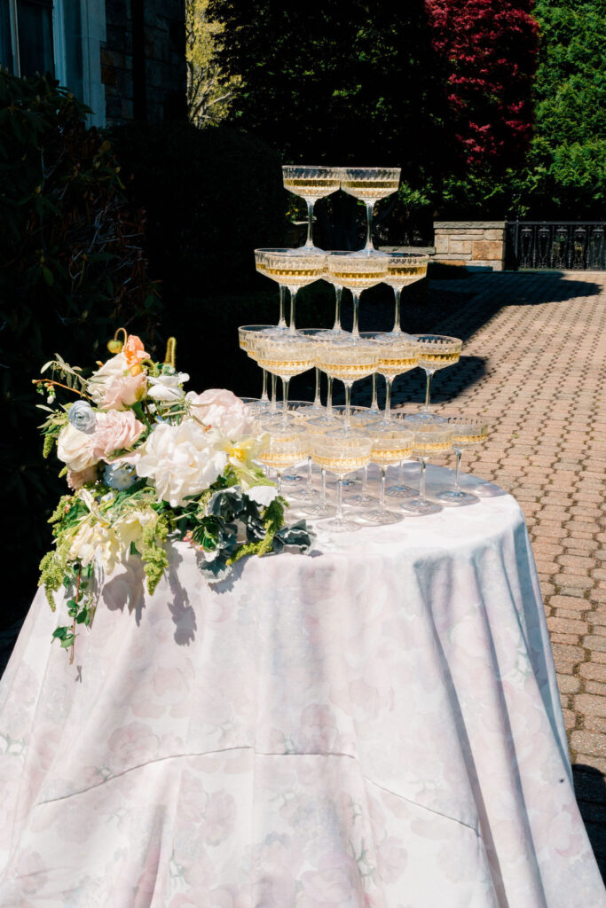 Shining Tides Weddings | Caprio Captured Photography Light and Airy Wedding Photography | Champagne tower on a table adorned with a floral arrangement and pastel cloth, set outdoors on a sunlit brick patio, conveying celebration.