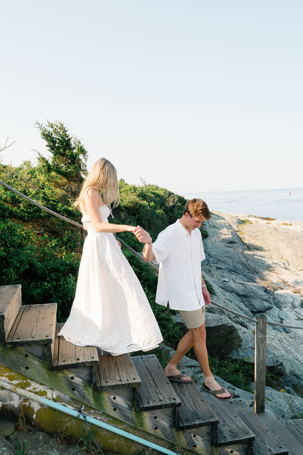 Newport Wedding Photographers | Caprio Captured Photography Light and Airy Wedding Photography | A couple descends wooden steps by the sea, holding hands. The woman wears a flowing white dress, and the man is in a white shirt and shorts. It's a serene, sunny day.