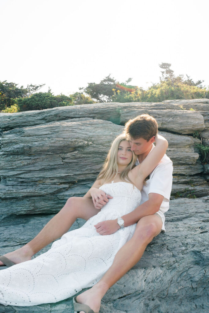 Newport Wedding Photographers | Caprio Captured Photography Light and Airy Wedding Photography | A couple in white summer clothes sits on rocks, embracing lovingly. The woman, in a long dress, leans back against the man. Sunlight highlights them.