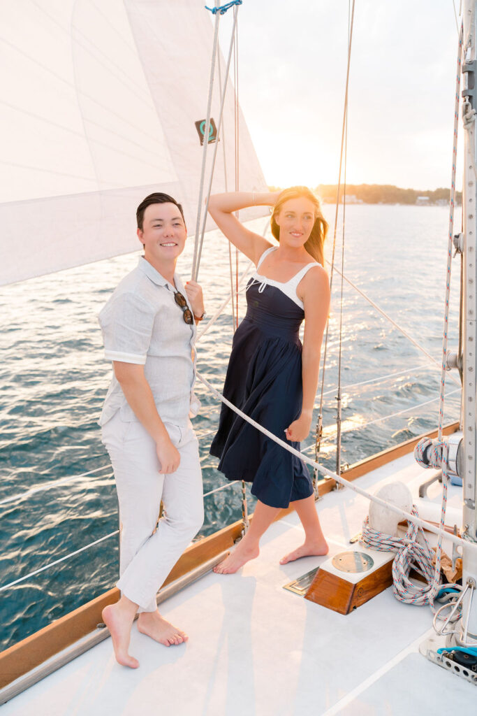 Newport Wedding Photographers | Caprio Captured Photography Light and Airy Wedding Photography | A couple enjoys a sunny day on a sailboat. The woman in a navy dress smiles, holding a sail rope. The man in a light outfit stands nearby. The ocean sparkles.