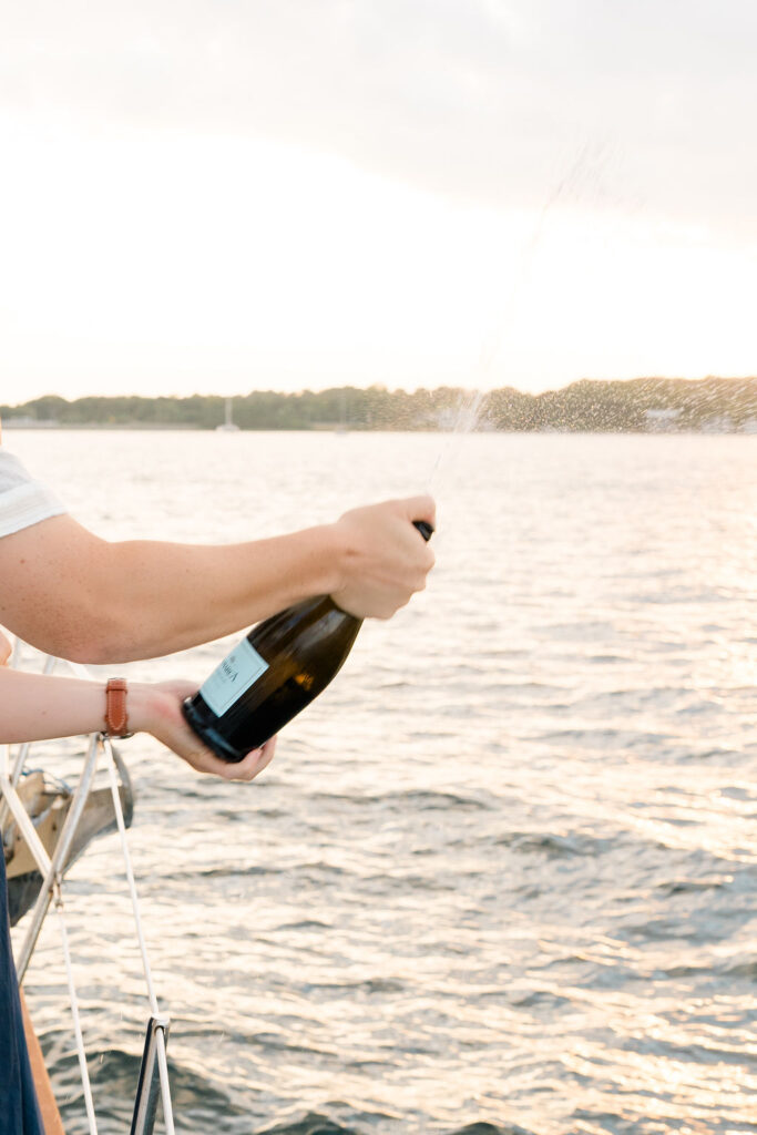 Shining Tides Weddings | Caprio Captured Photography Light and Airy Wedding Photography | A person pops open a champagne bottle on a boat, spraying bubbly liquid over a sunlit, serene body of water, evoking celebration and joy.
