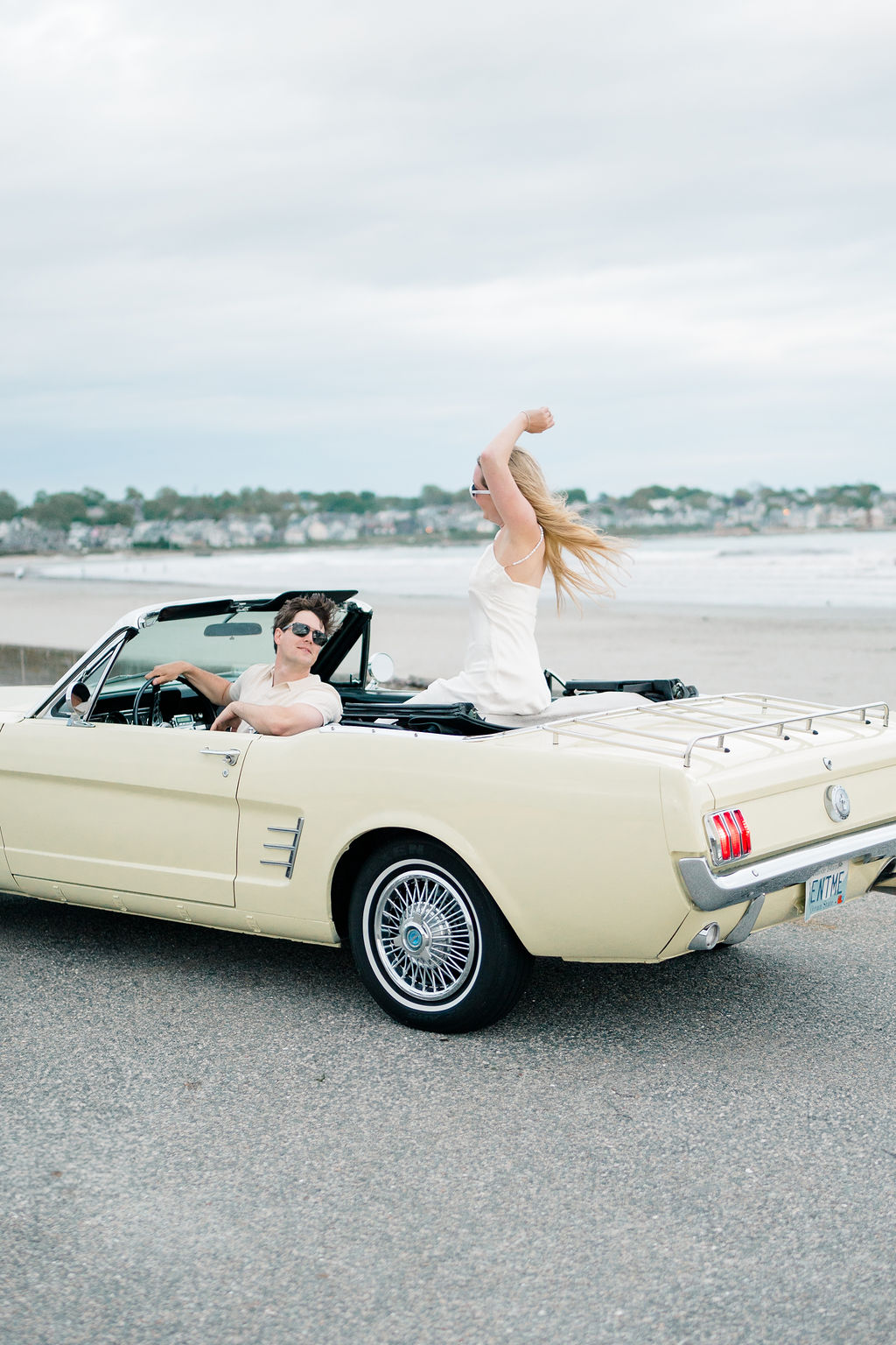Waterfront Wedding Venues Rhode Island | Caprio Captured Photography Light and Airy Wedding Photography | A couple enjoys a ride in a vintage cream convertible by the beach. The woman stands, arms raised joyfully, while the man drives, both smiling broadly.